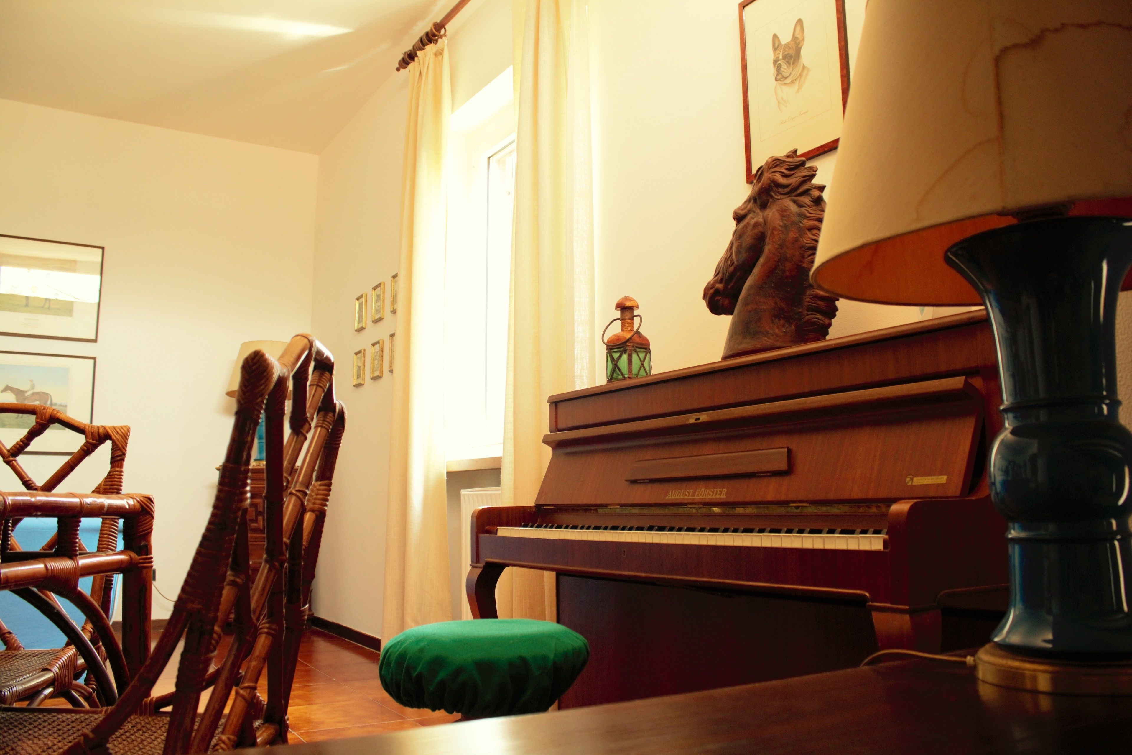 A room with a wooden piano, a green cushioned bench, a tall lamp, and framed pictures on the wall.