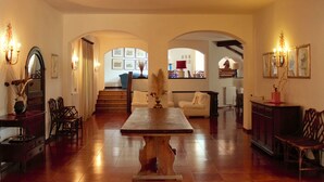 A spacious living area with a wooden dining table, multiple chairs, and a sofa. There are archways leading to other rooms, and the space is well-lit with wall-mounted lights.