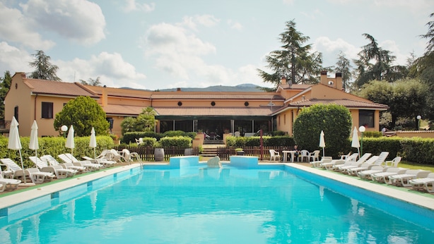 A swimming pool with lounge chairs and umbrellas, surrounded by a building with a tiled roof and a view of hills in the background.