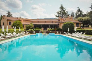 A swimming pool with lounge chairs and umbrellas, surrounded by a building with a tiled roof and a view of hills in the background. - La Ripa (Scandriglia)