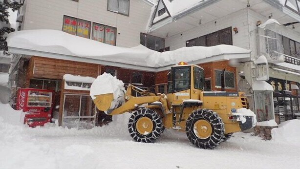 A yellow snowplow in front of a building with a Coca-Cola vending machine. - Hakuba Cortina Backpackers - Hostel (Otari)