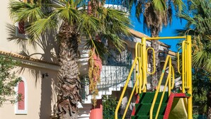 Un parque infantil con un tobogán amarillo y escalones verdes, rodeado de palmeras y un edificio con puerta roja.