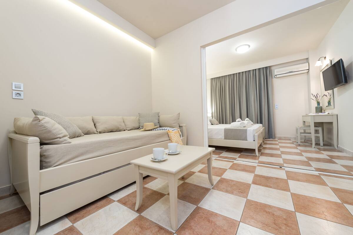 A modern hotel room with a sofa, a small table with cups, a bed, and a TV mounted on the wall.