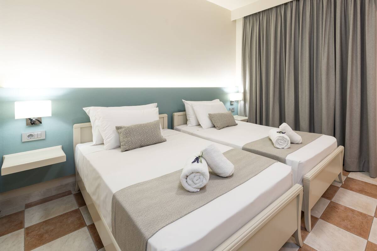 A hotel room with two beds, a small bedside table with a lamp, and a large window with curtains.