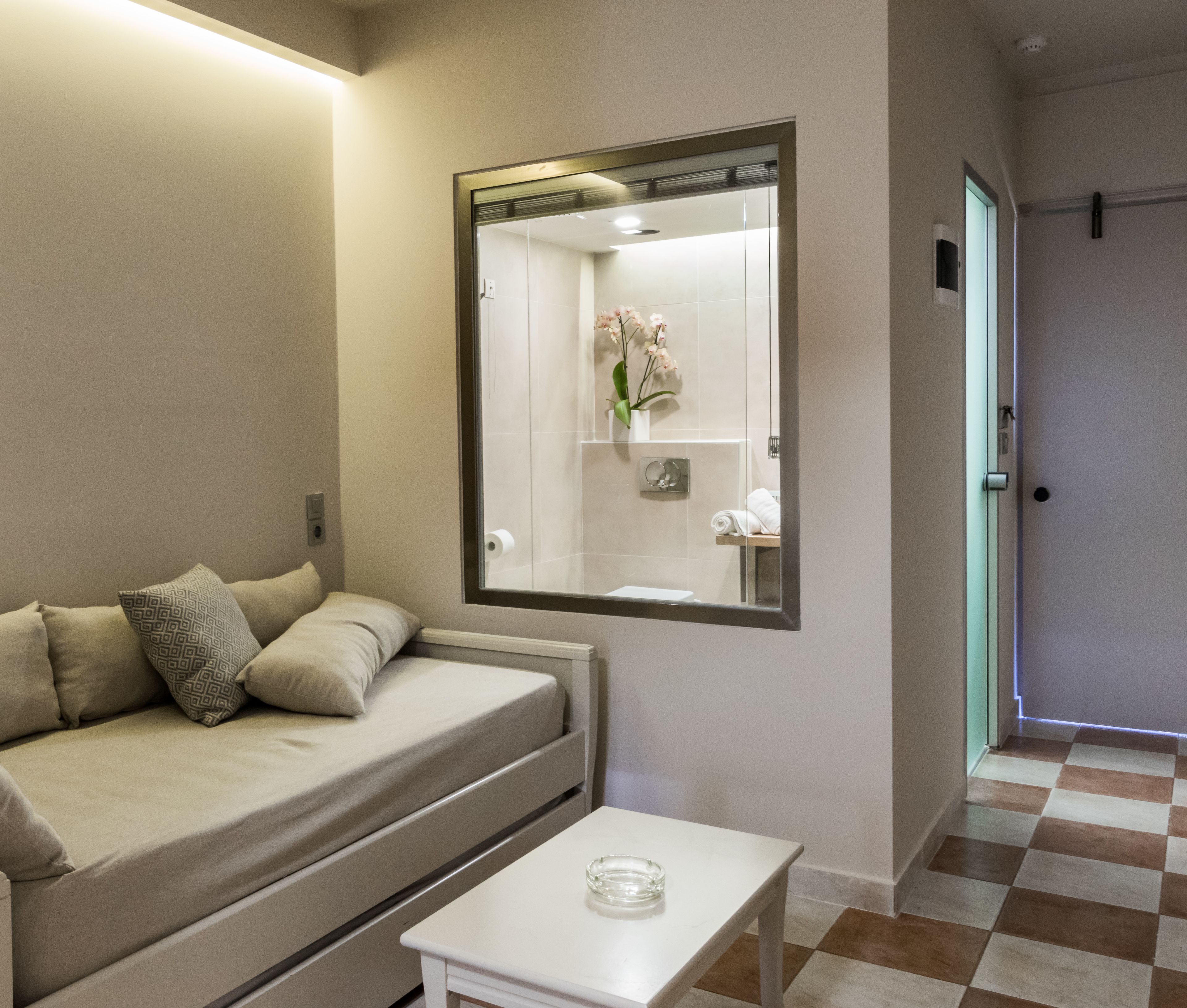 A compact room with a sofa, a small table, and a mirror reflecting a bathroom with a sink and a towel rack.