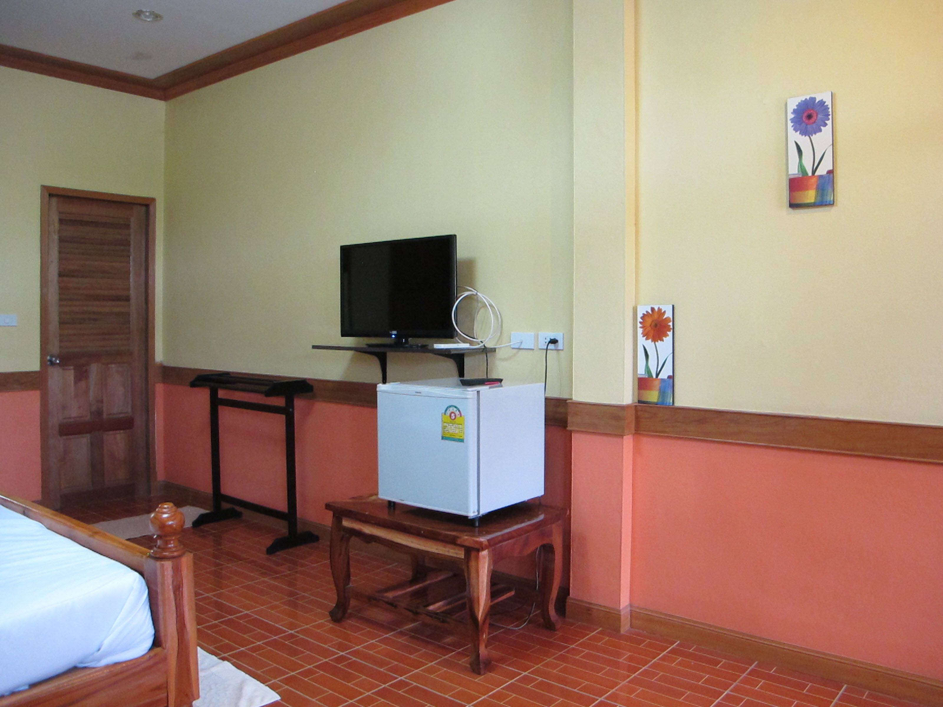 Una habitación con una cama, un escritorio, un televisor sobre un soporte, un refrigerador pequeño y una puerta.
