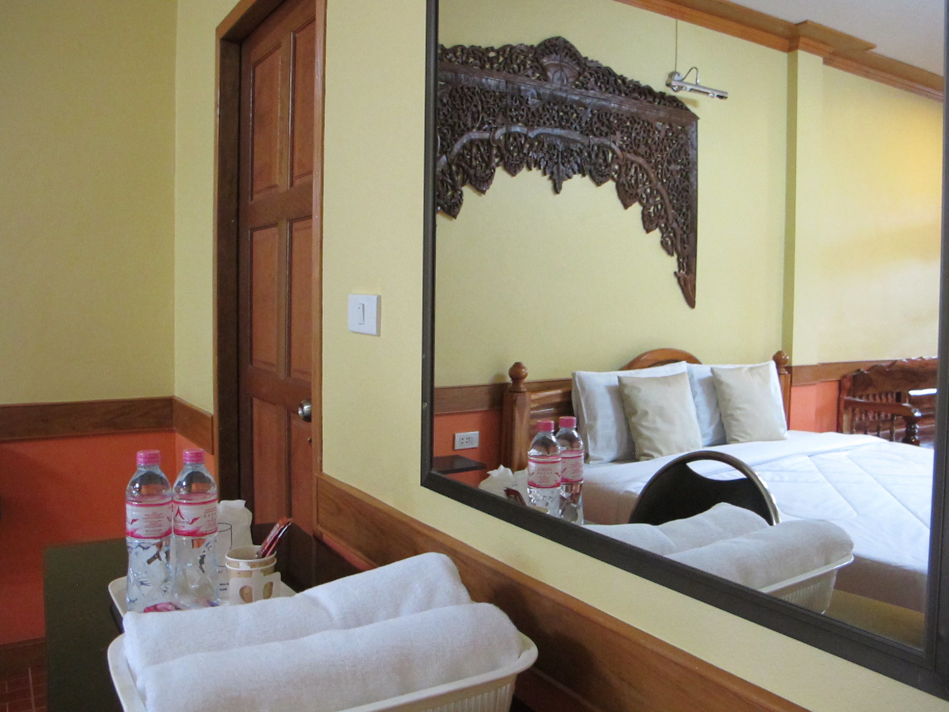 Une chambre d’hôtel avec une porte en bois, un grand miroir qui reflète le lit et une tête de lit en bois, ainsi qu’une petite table sur laquelle se trouvent des bouteilles et une boîte de mouchoirs.