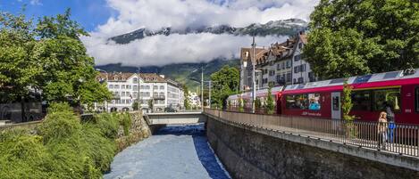 A river running through a town with buildings on either side, a bridge, and mountains in the background.