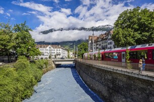 A river running through a town with buildings on either side, a bridge, and mountains in the background.