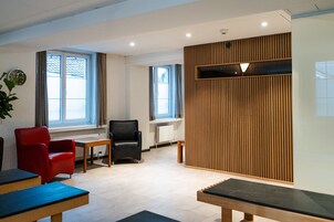 Un área de espera moderna con una silla roja, una silla negra y un banco de madera. Hay ventanas con persianas, un radiador y una estantería en la pared.