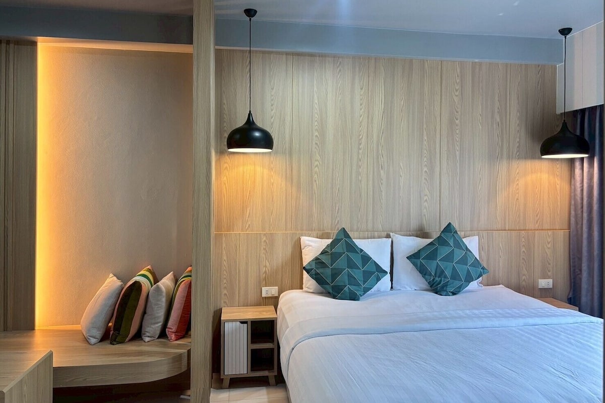 A modern bedroom with a wooden headboard, a bed with white bedding, two geometric-patterned pillows, a nightstand, and a wall-mounted lamp.