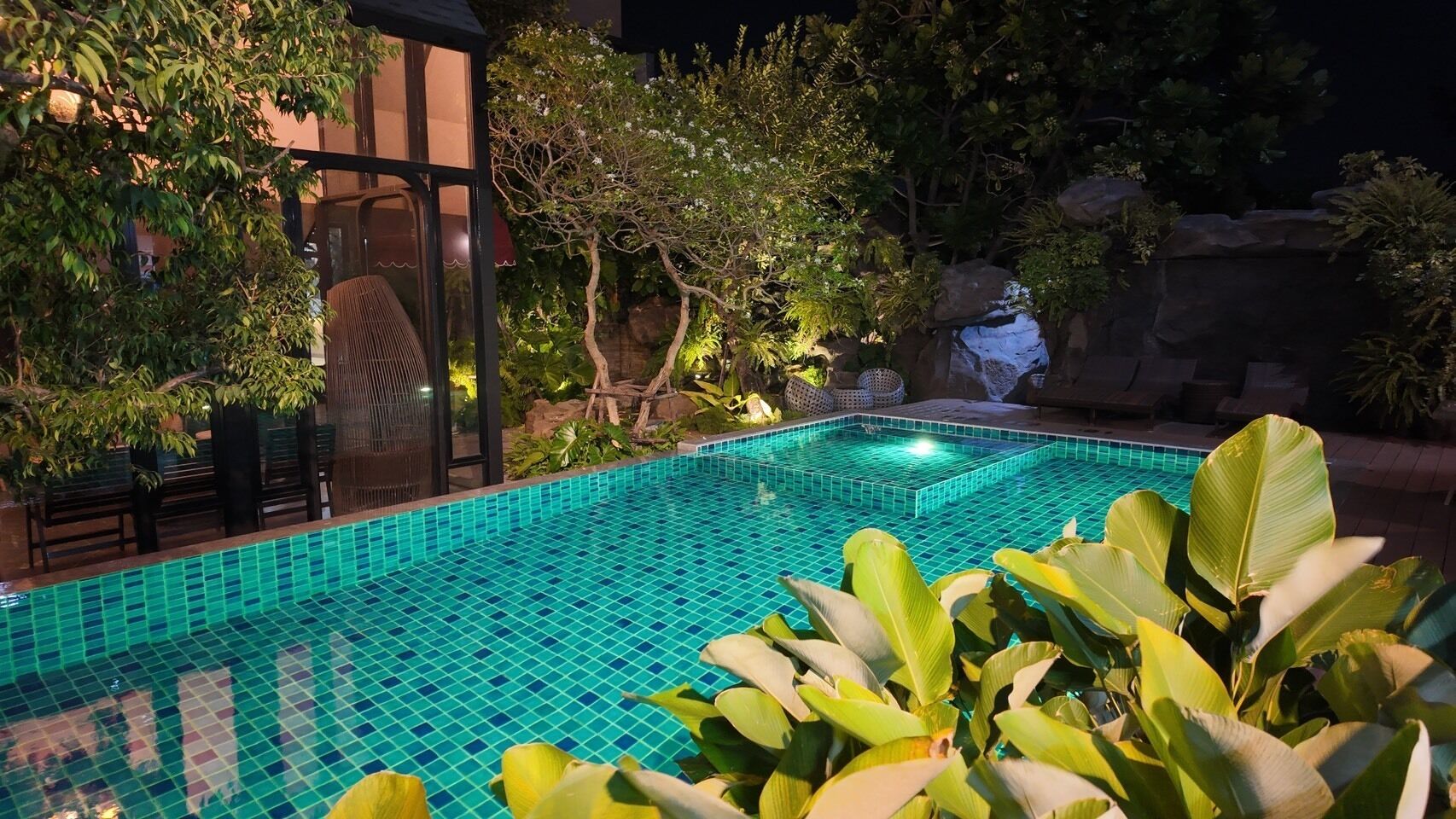 Una piscina con borde de azulejos, rodeada de exuberante vegetación y una formación rocosa.