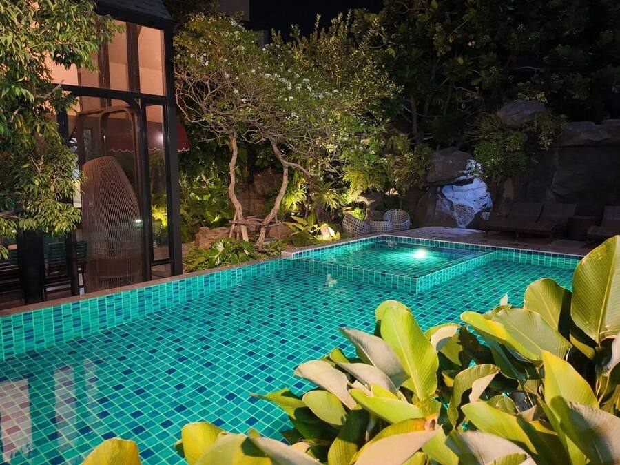 Una piscina con borde de azulejos, rodeada de exuberante vegetación y una formación rocosa.