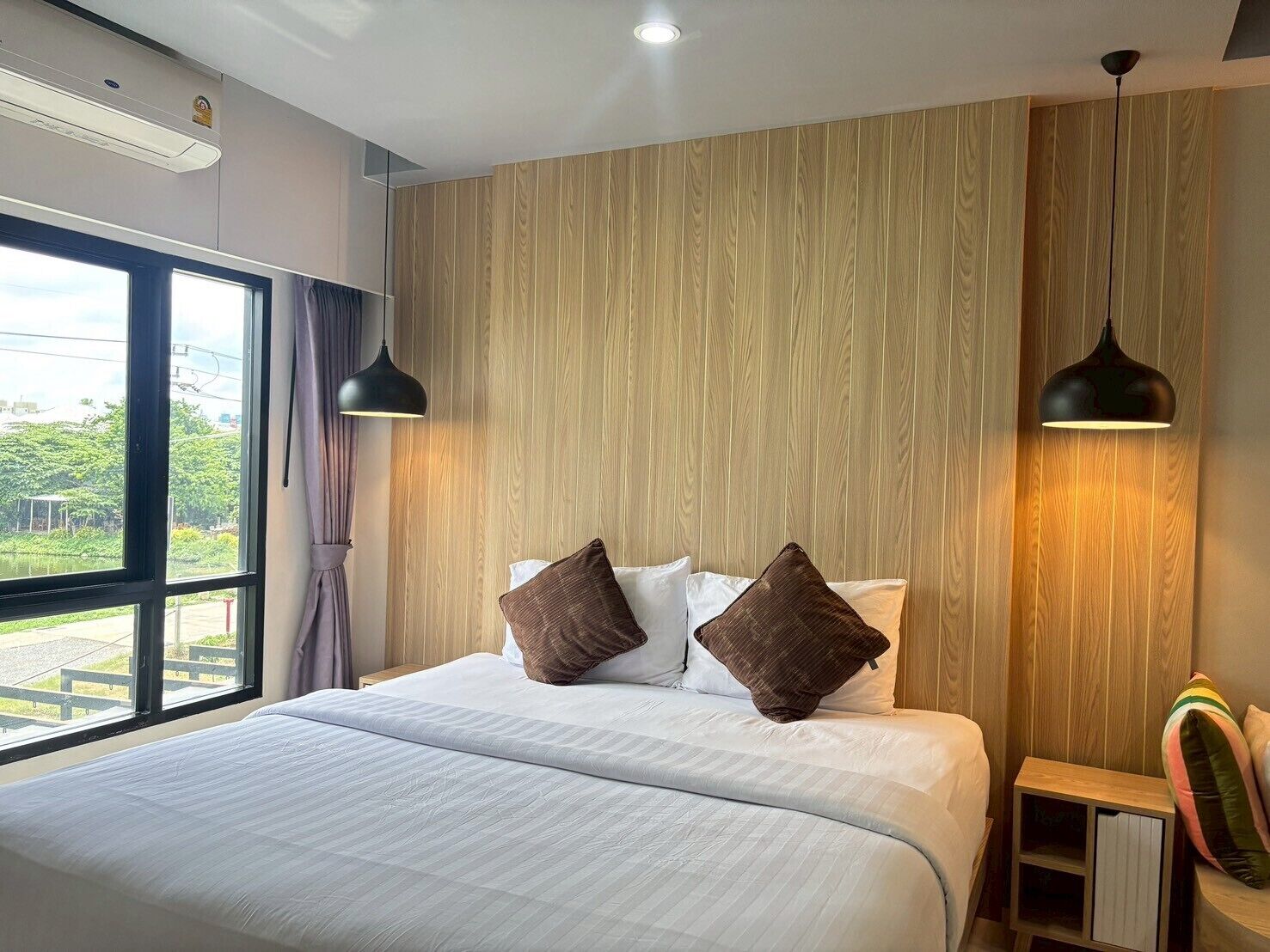 A modern hotel room with a large bed, wooden panel walls, and a window with a view of greenery.