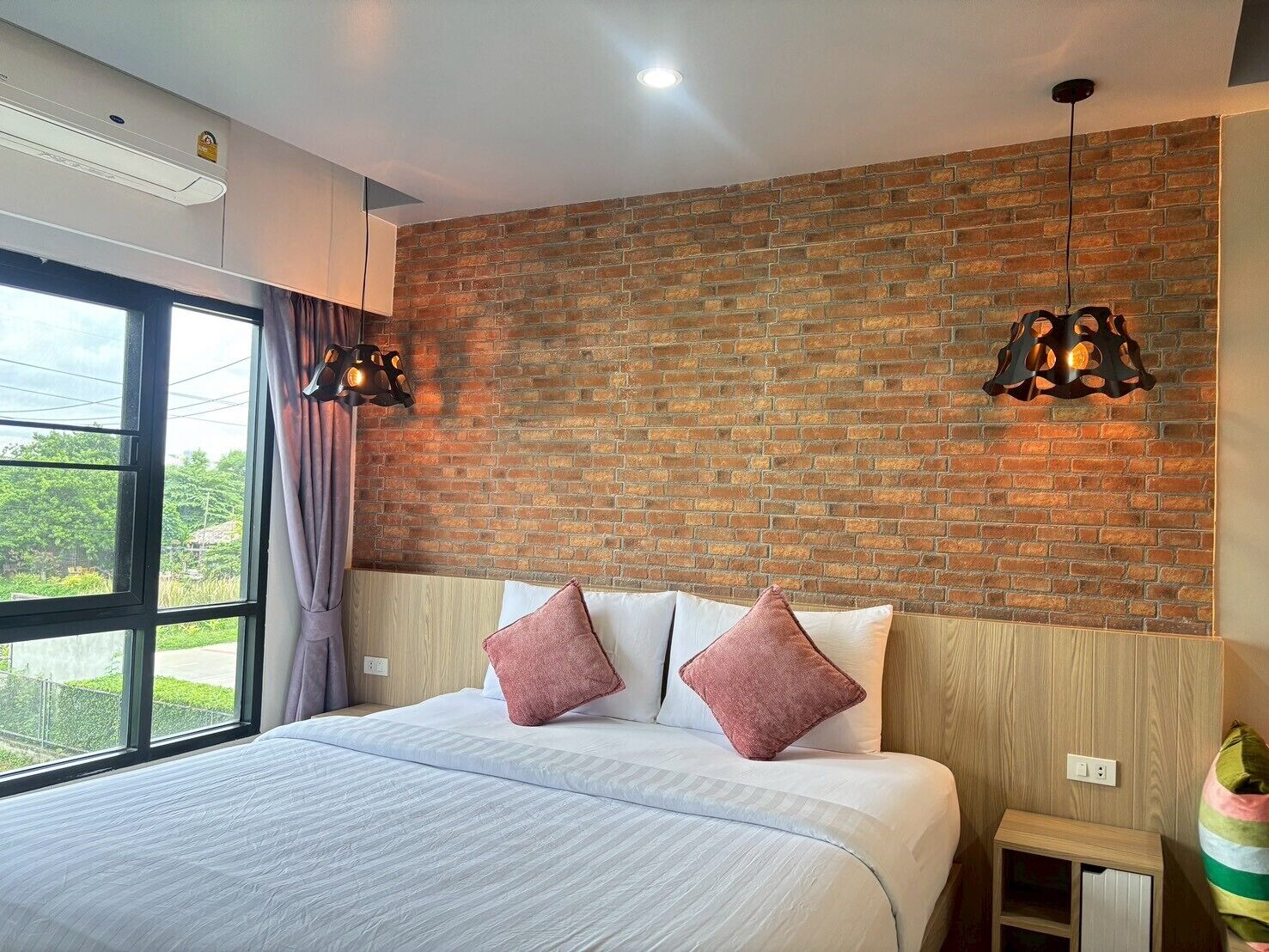 Un dormitorio con pared de ladrillo, una cama grande con almohadas blancas y rosas, dos lámparas colgantes y una ventana con cortinas.