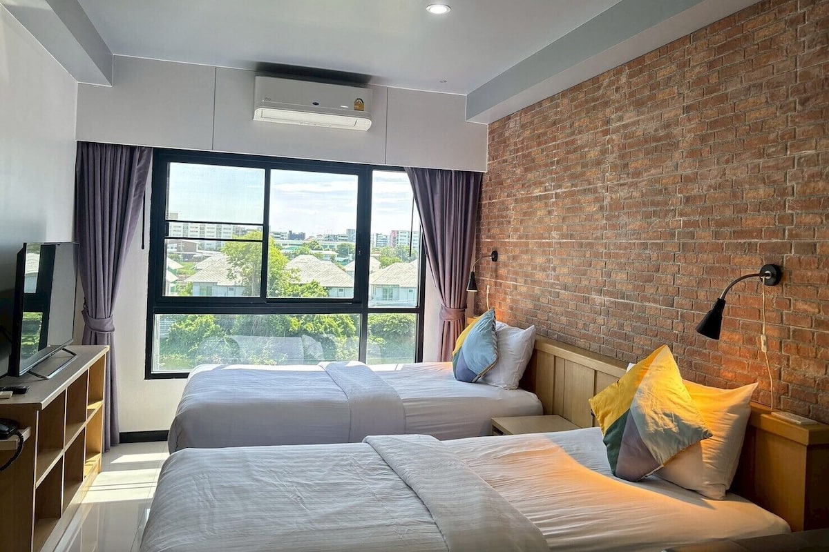 A hotel room with two beds, a brick wall, a large window, and a TV.