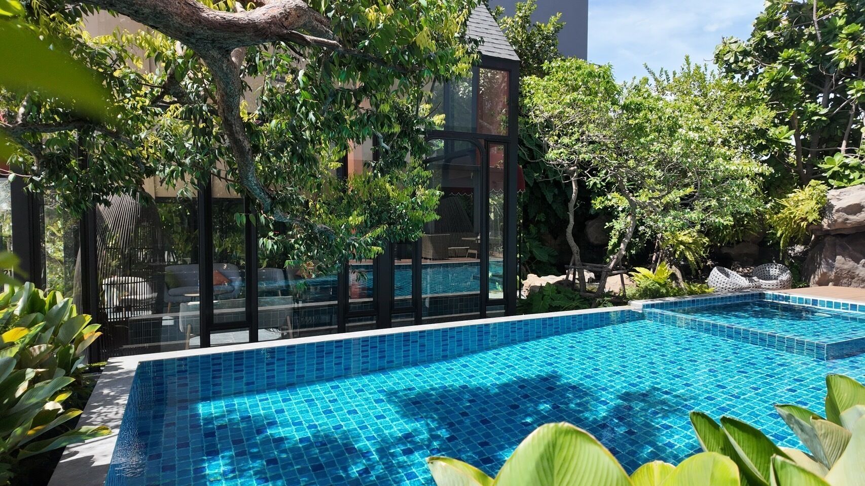Una piscina rectangular con azulejos azules, rodeada de exuberante vegetación y una estructura con cerramiento de vidrio.