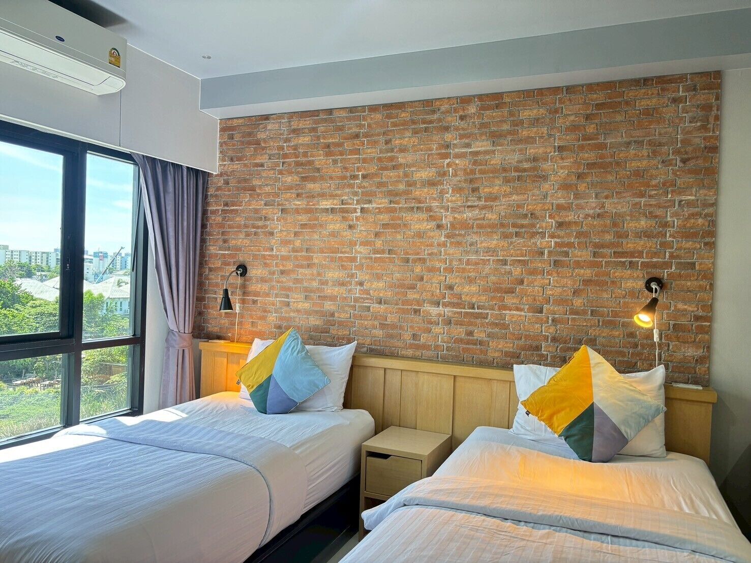 Een hotelkamer met twee bedden, een bakstenen muur, een raam met gordijnen en uitzicht op de stad.