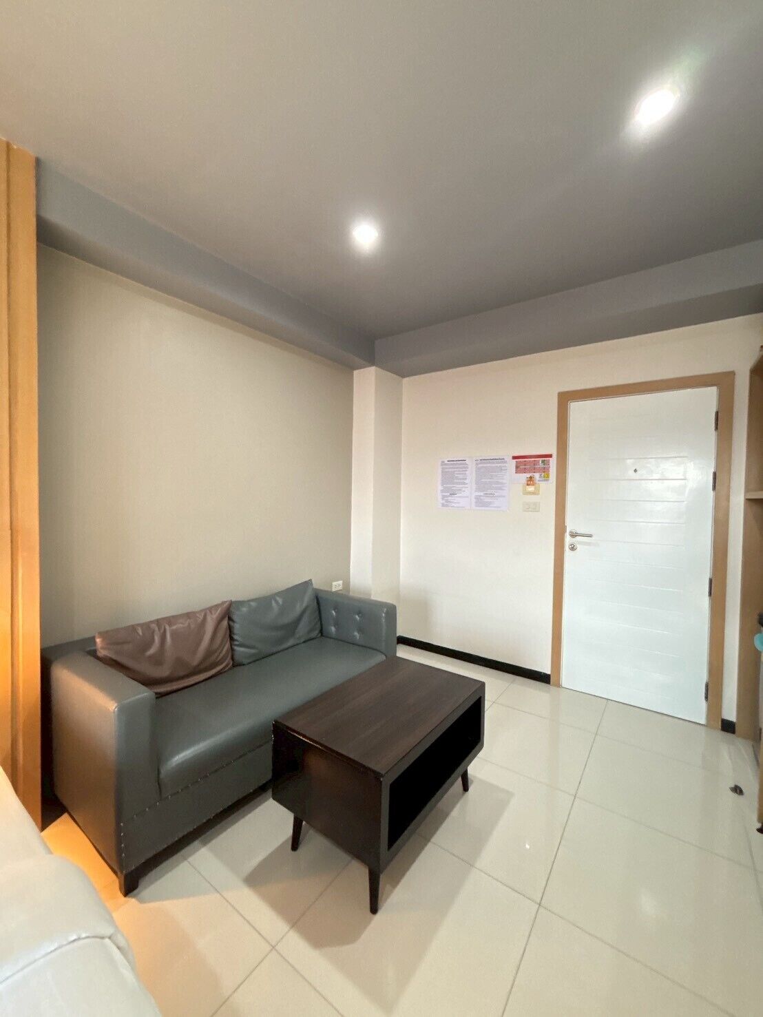 A living room with a grey sofa, a dark wooden coffee table, a white door, and a bulletin board on the wall.
