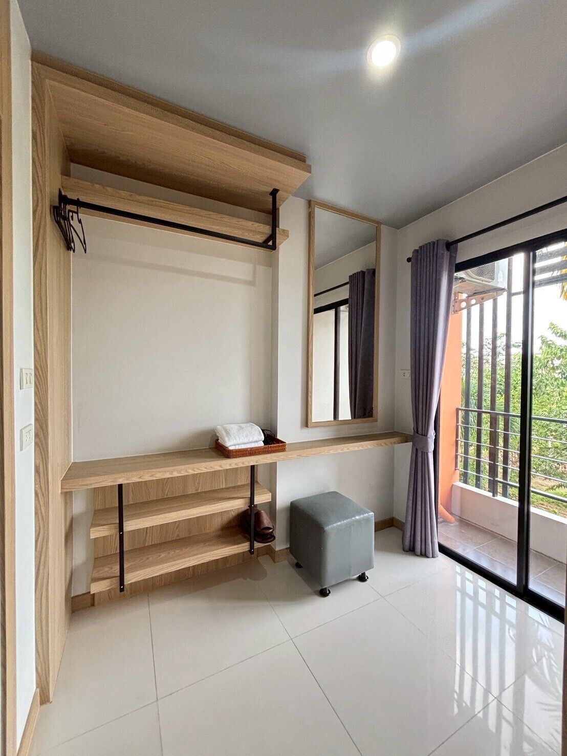 A modern room with a wooden bench, a small ottoman, a mirror, and a view through a window with curtains.