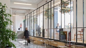 A spacious indoor area with large windows, seating arrangements, and a staircase leading to an upper level.