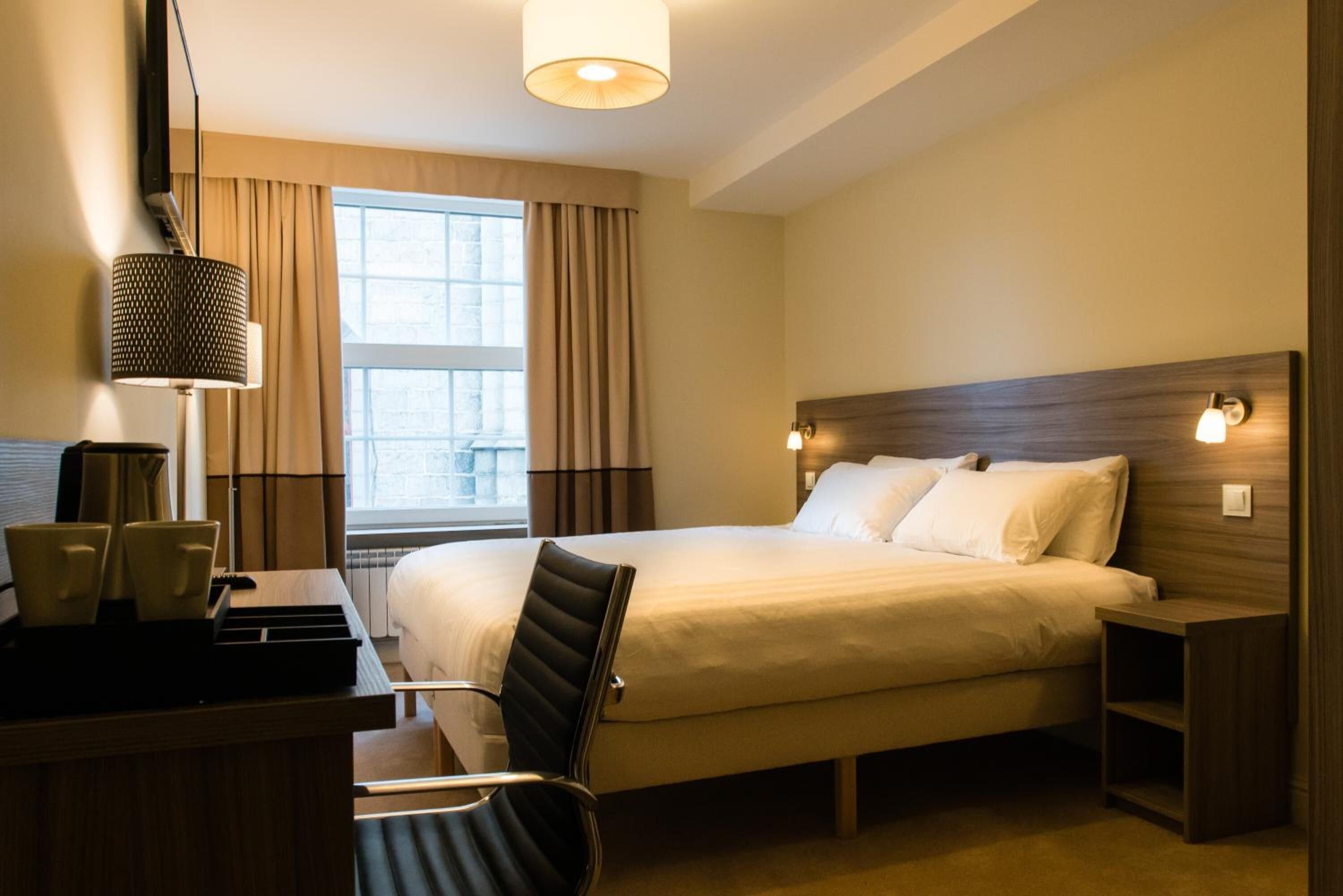 A hotel room with a large bed, a desk with a chair, a lamp, and a window with curtains.
