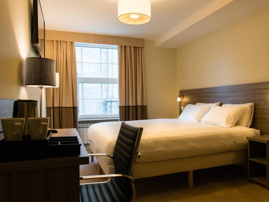 A hotel room with a large bed, a desk with a chair, a lamp, and a window with curtains.