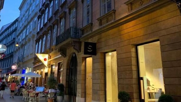 A street view of a building with a large 'S' sign, indicating a hotel or similar establishment. There are outdoor seating areas with tables and chairs, suggesting a cafe or restaurant on the ground floor. - KH Apartments (Vienna)
