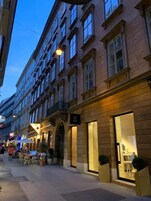 Vista da rua de um edifício com um grande letreiro em formato de 'S', indicando um hotel ou estabelecimento similar. Possui áreas de estar ao ar livre com mesas e cadeiras, sugerindo um café ou restaurante no andar térreo.