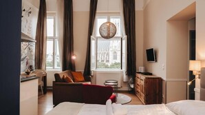 A hotel room with a bed, a red armchair, a desk, a television, and a view of a building through the window. - KH Apartments (Vienna)