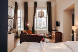 Quarto de hotel com cama, poltrona vermelha, escrivaninha, televisão e vista para um edifício pela janela.