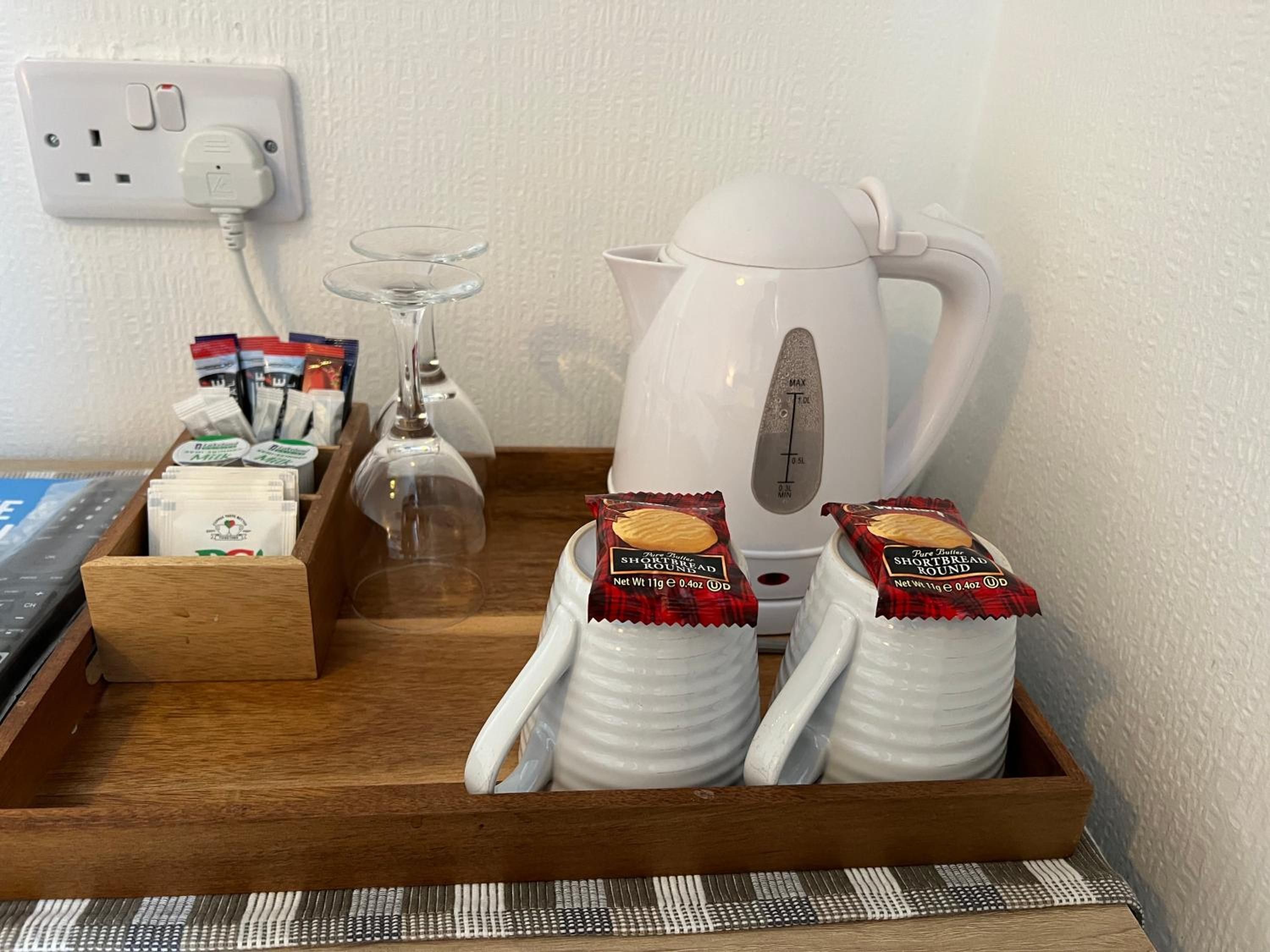 A breakfast tray with a white kettle, two glass tumblers, a wooden tray, and packets of shortbread.