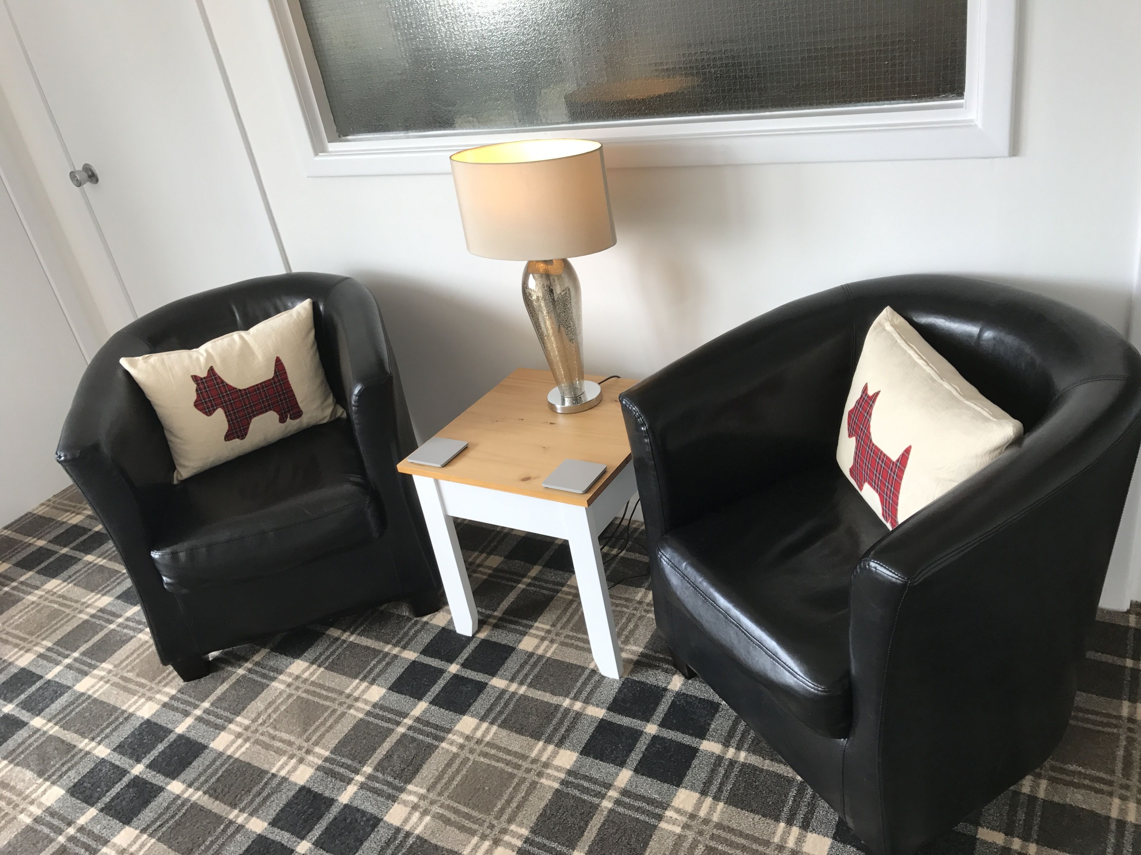 Dos sillones de cuero negro con cojines estampados de pata de gallo, una mesita de madera con una lámpara, y un piso a cuadros.