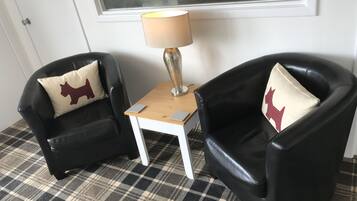 Dos sillones de cuero negro con cojines estampados de pata de gallo, una mesita de madera con una lámpara, y un piso a cuadros.