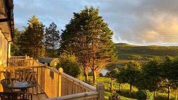 Una terraza de madera con mobiliario exterior, con vistas a un paisaje con césped, árboles y una colina a lo lejos.