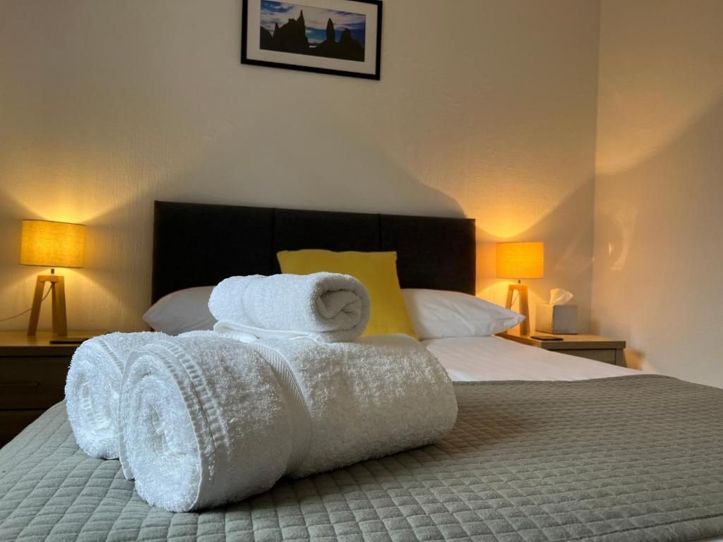 A neatly made bed with rolled white towels, a quilted grey bedspread, and a yellow pillow. There are two bedside lamps on wooden nightstands.