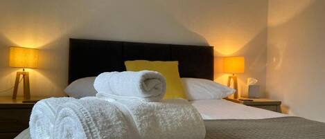 A neatly made bed with rolled white towels, a quilted grey bedspread, and a yellow pillow. There are two bedside lamps on wooden nightstands.