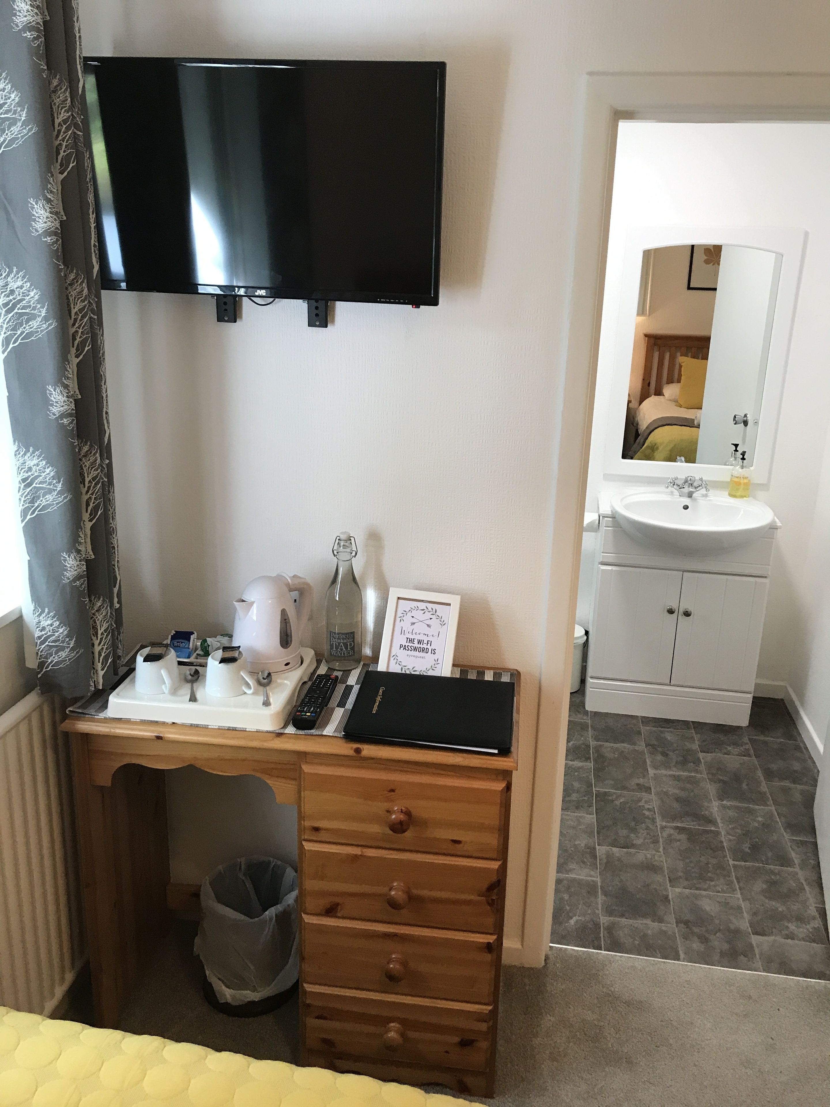 Une chambre d’hôtel avec un téléviseur fixé au mur, un bureau en bois sur lequel se trouve un plateau avec une bouilloire, une bouteille d’eau et une pancarte indiquant le mot de passe du Wi-Fi. La salle de bain est équipée d’un lavabo blanc et d’un miroir.