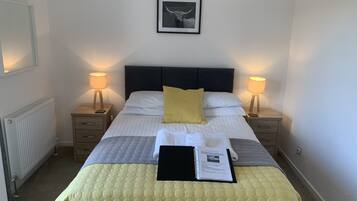 Una cama bien tendida con un edredón amarillo, dos lámparas de noche, un cuadro enmarcado en la pared y una mesita de noche con un libro y un teléfono.