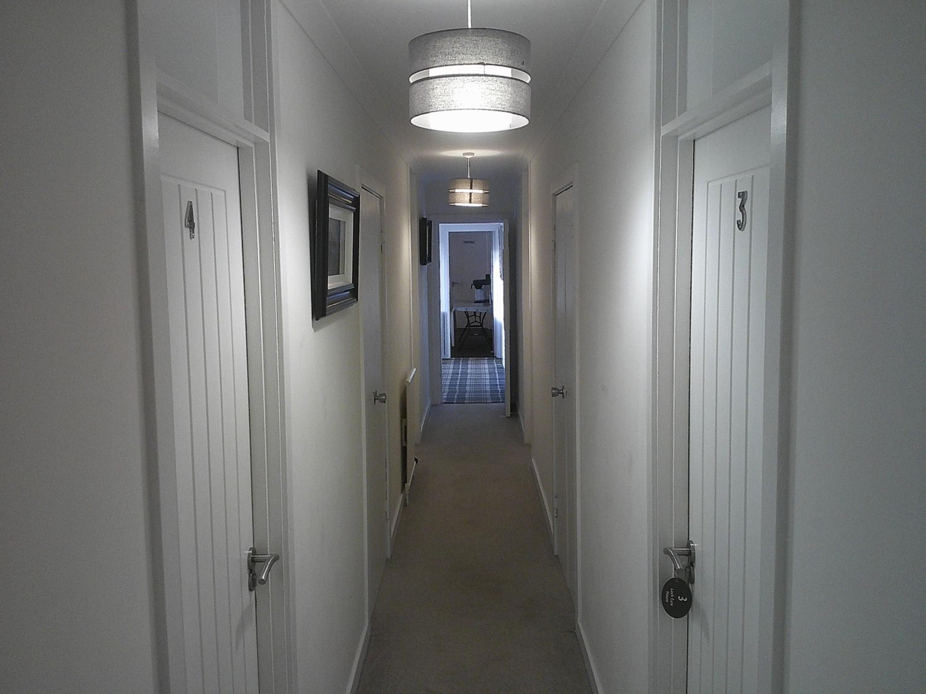 A narrow hallway with white doors, a hanging light fixture, and a checkered floor leading to a room with a bed and a chair.