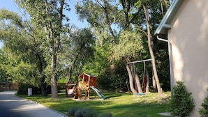 Un terrain de jeux avec un toboggan et des balançoires, entouré d’arbres et bordé d’un bâtiment.