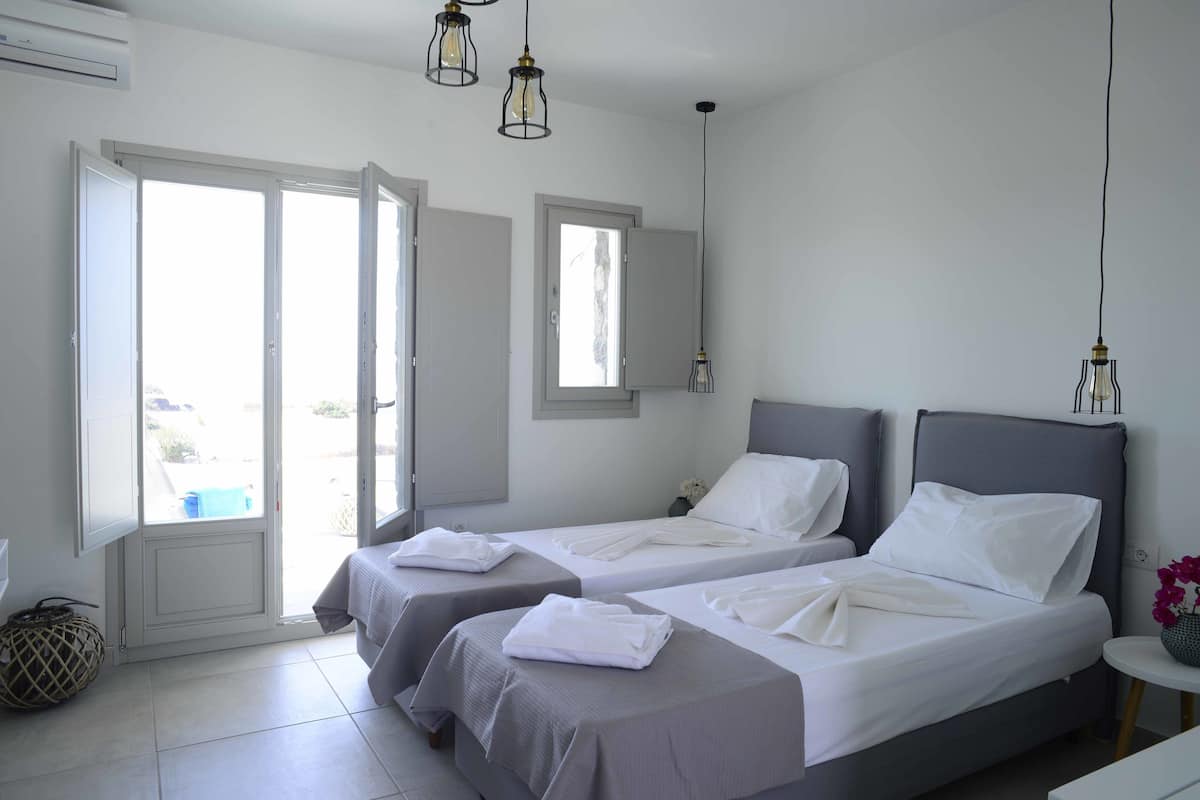 Habitación de hotel con dos camas, una mesita, una silla, un ventanal y una puerta que da a un espacio al aire libre.