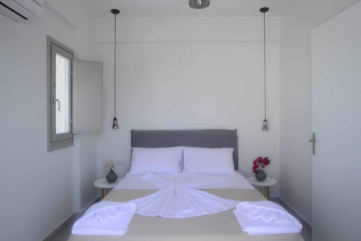 Una cama bien hecha con sábanas blancas, dos mesitas de noche con jarrones y una ventana con vistas.