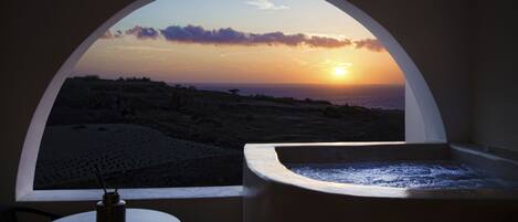 A room with a view of a sunset over the ocean, a bathtub, and a small table with a plant.
