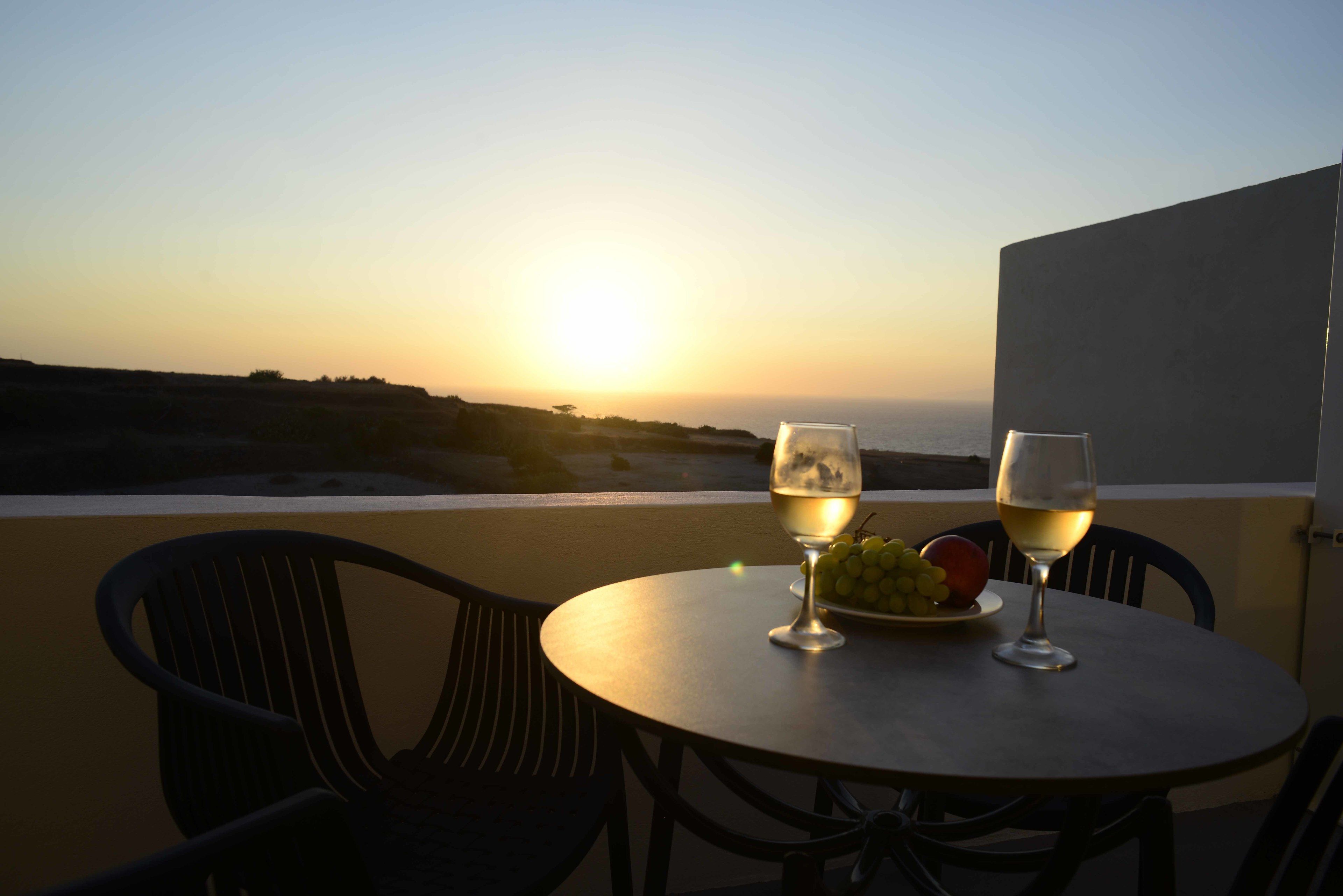 A table with two glasses of white wine and a plate of grapes and an apple, overlooking a sunset over the sea.