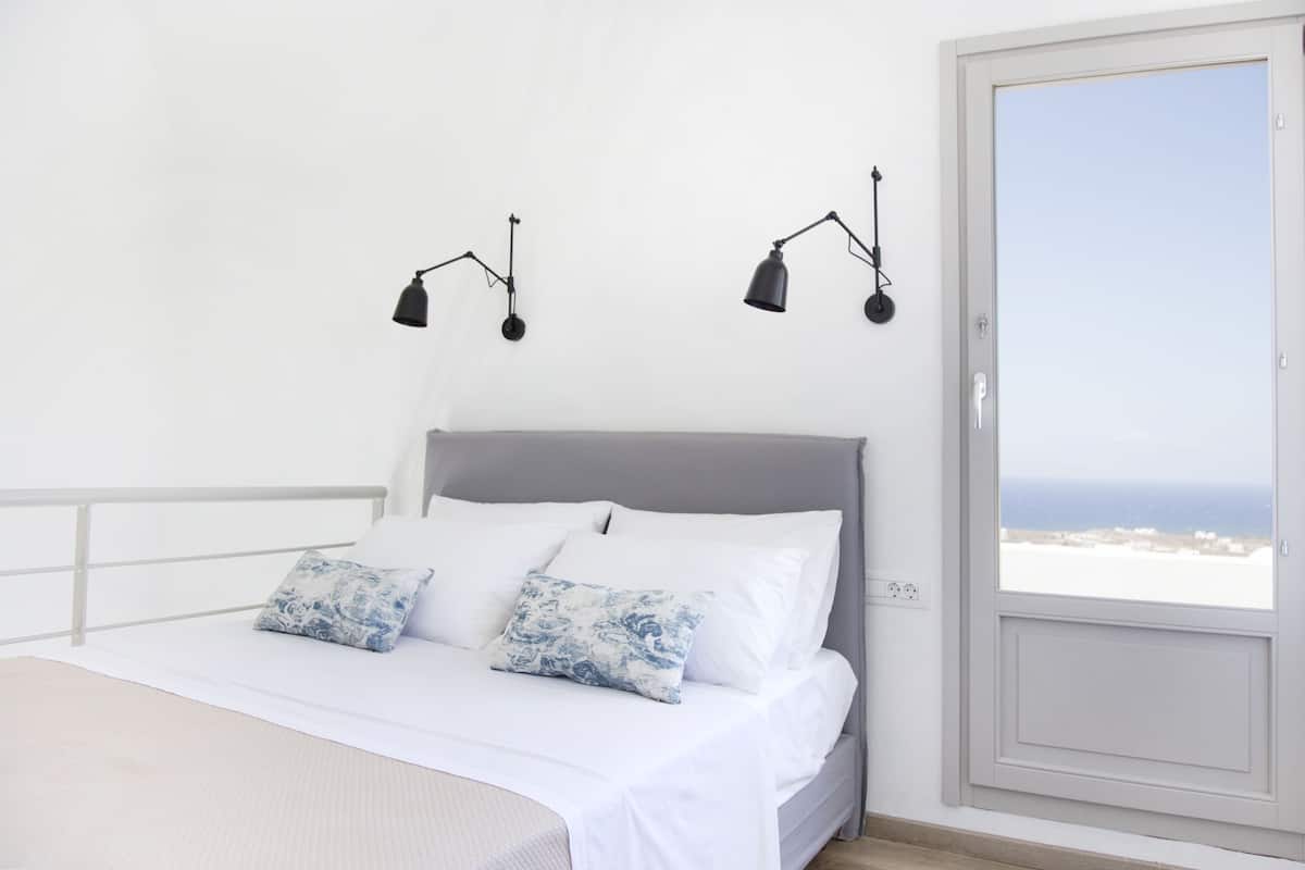 Un dormitorio con una cama, ropa de cama blanca, dos lámparas fijadas a la pared y vista al mar a través de una puerta.
