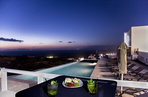 Une vue sur la piscine avec un coucher de soleil sur l’océan, une table garnie de nourriture et de boissons, et des chaises longues.