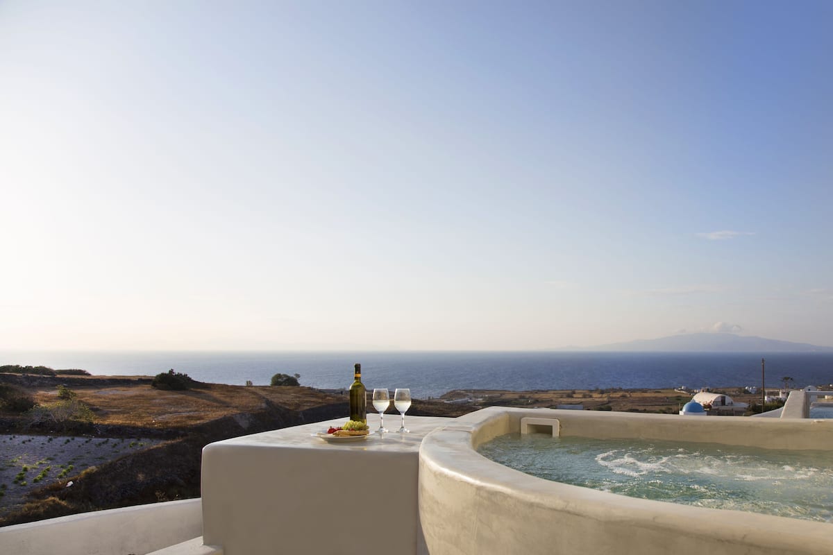 Una terraza con jacuzzi, una botella de vino y una copa sobre una mesa, con vista a un paisaje costero y una montaña en la distancia.