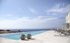A poolside area with lounge chairs and two wicker baskets, overlooking a clear blue sea and a distant coastline. - Aplai Dome (Santorini)