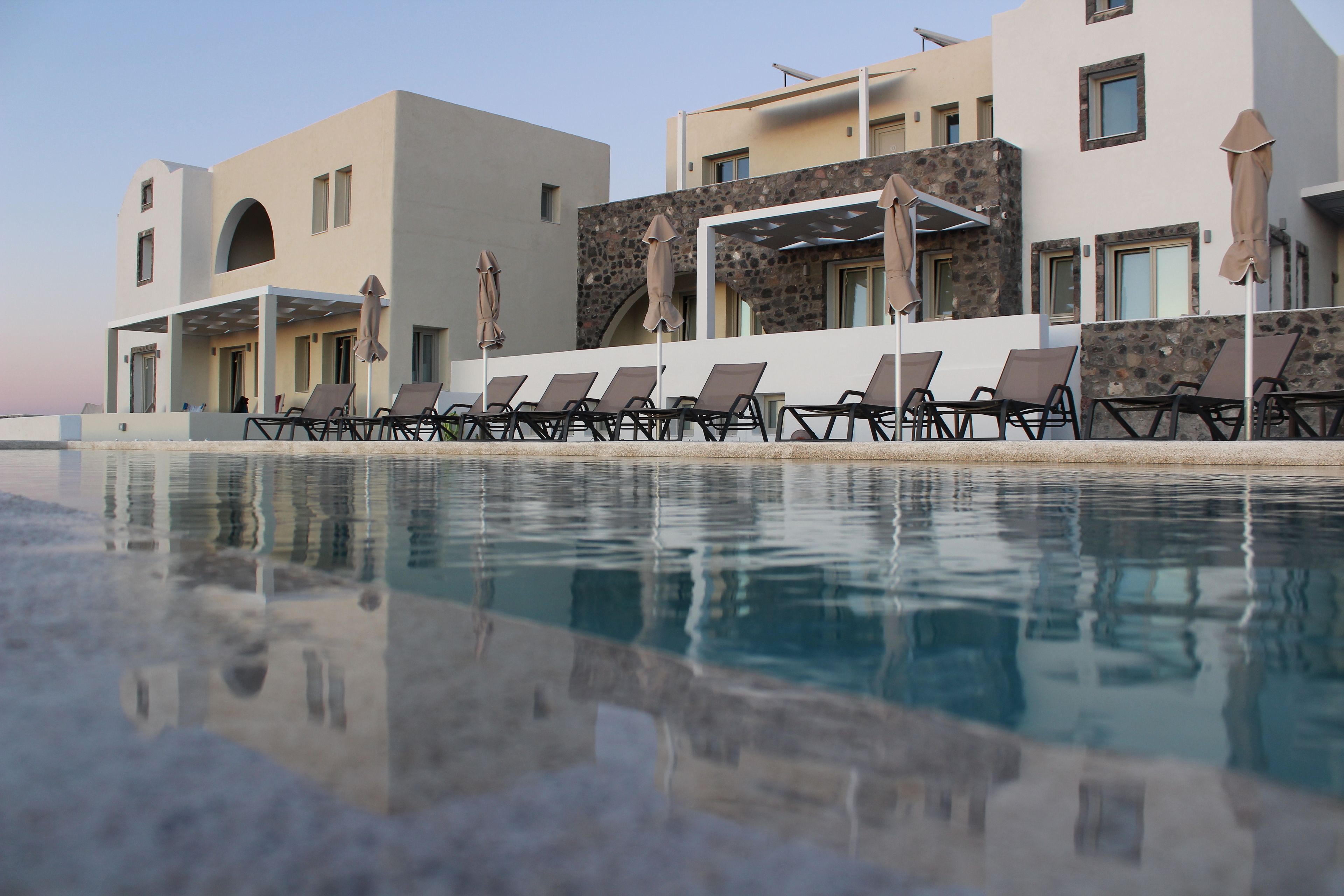 Un área junto a la piscina con sillones de descanso y sombrillas, piscina cristalina que refleja los edificios, y un edificio moderno con detalles de piedra.